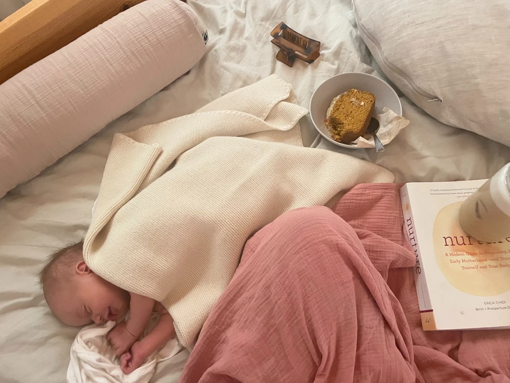ein lächelndes Baby im Bett gemeinsam mit einem Buch, Flasche und Haarspange aus der Perspektive der Mama, schöne pastellige Stoffe und Bettwäsche, ein glücklicher Moment, reguliertes Baby